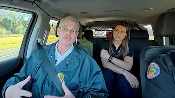 Hombre con gafas y chaqueta oscura en coche gesticulando. Mujer a su lado atenta. Pasajero trasero. Chaqueta con parche de Patrulla de Carreteras de Florida