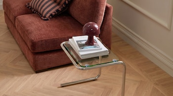 Un sofá de pana marrón rojizo, una mesa auxiliar de cristal y cromo con libros y una lámpara de hongo burdeos, y un suelo de madera en espiga.