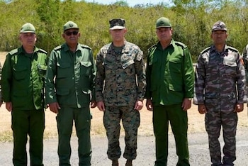 Cinco hombres con uniformes militares de pie en fila frente a un fondo boscoso. Un soldado central lleva uniforme de camuflaje, los demás visten uniformes verdes o estampados
