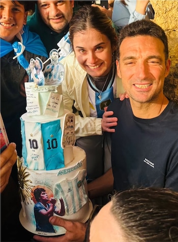 Lionel Scaloni y una mujer sonríen, con otras personas alrededor y una torta de tres pisos decorada con imágenes de futbolistas y camisetas argentinas
