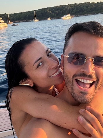 Primer plano de una mujer con el pelo mojado abrazando por detrás a un hombre que lleva gafas de sol, ambos sonríen en un barco con el mar y otras embarcaciones de fondo