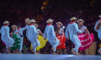 Bailarines de folclore con trajes tradicionales y sombreros en un escenario. Hombres con ropa blanca, mujeres con faldas coloridas. Público de fondo