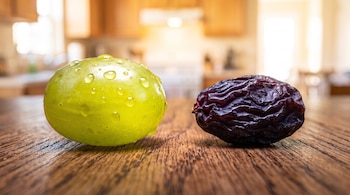 Una uva verde con gotas de agua y una uva pasa arrugada colocadas sobre una mesa de madera en una cocina.
