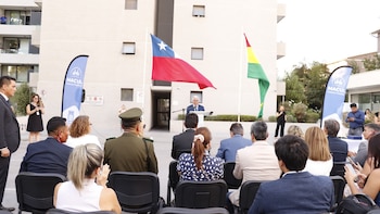 Un hombre canoso con traje azul habla desde un podio al aire libre frente a una multitud, con las banderas de Chile y Bolivia ondeando a su lado y edificios de fondo
