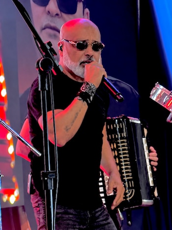Hombre calvo con barba canosa, gafas de sol y camiseta negra canta en un micrófono junto a un acordeonista