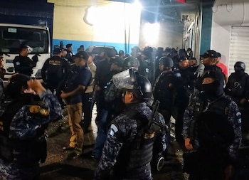 Grupo de agentes policiales y de seguridad con uniformes camuflados, chalecos, algunos con cascos y escudos, de noche, junto a un camión blanco y un edificio