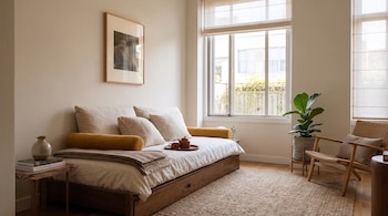 Sofá cama con cojines claros y amarillos, bandeja de té, cuadro en la pared, alfombra de yute, planta en maceta y un sillón junto a la ventana.