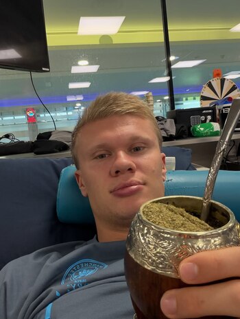 Erling Haaland recostado en un sillón azul sostiene en primer plano un mate con yerba y bombilla metálica frente a una pared de vidrio