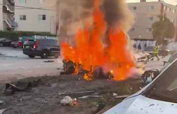 Restos de un coche ardiendo intensamente con grandes llamas y humo; al fondo, un vehículo de la policía de Miami-Dade con luces intermitentes y escombros