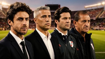 Cuatro hombres vestidos con trajes oscuros y prendas deportivas permanecen alineados frente al césped de un estadio de fútbol