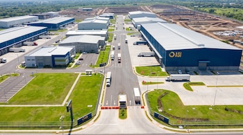 Vista aérea panorámica de un parque industrial moderno en El Salvador, con naves azul profundo y gris, camiones en calles, áreas verdes y construcción al fondo.