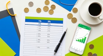 Vista cenital de una mesa de trabajo con hoja de cálculo impresa, un bolígrafo, monedas, taza de café y un smartphone mostrando una aplicación de finanzas.