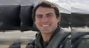 Retrato de un hombre sonriente con indumentaria de vuelo frente a una aeronave militar