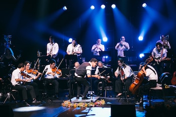 Conjunto de músicos en un escenario oscuro con fuertes luces azules, tocando violines, saxofones, trompetas y guitarra. Hay flores en el suelo