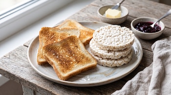Plato con tostadas de pan y tortitas de arroz inflado, acompañado de recipientes con mantequilla y mermelada sobre una mesa de madera.