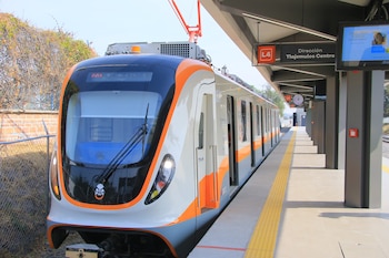 Tren blanco, naranja y gris estacionado en una plataforma. Sus puertas están abiertas. Un cartel rojo indica 'L4' y otro oscuro 'Dirección Tlajomulco Centro'. Se aprecian vías