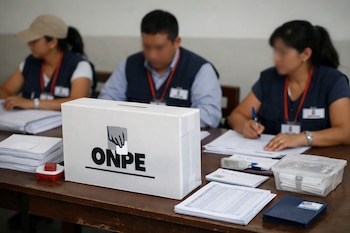 Urna blanca con logo de ONPE, material electoral y tres personas con chalecos azules y credenciales sentadas en una mesa de madera.