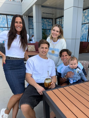 Eva Bargiela, Carolina Baldini, dos hombres y un bebé posan en un patio; una mujer lleva un suéter con la bandera argentina y un hombre sostiene un mate