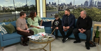 Cuatro personas (dos hombres, una mujer, un niño) sentadas en un sofá azul en un estudio de TV. Una mesa de centro está enfrente. Un paisaje urbano se ve al fondo