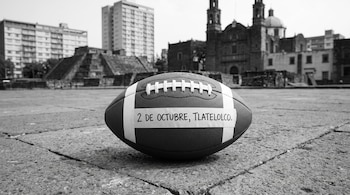 Un balón de fútbol americano con una cinta que dice 2 DE OCTUBRE, TLATELOLCO sobre el suelo de piedra de una plaza con una pirámide e iglesia.