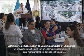 Hombre en podio con micrófono frente a banderas de Guatemala. Un grupo de asistentes lo observa frente a una pantalla con imágenes de videovigilancia de calles.