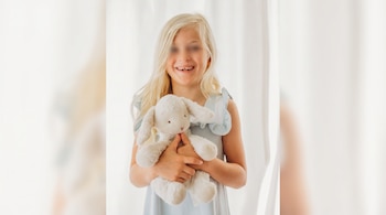 Niña rubia con vestido claro y rostro con ojos difuminados sostiene un conejo de peluche blanco frente a una cortina.