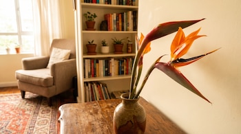Dos flores ave del paraíso de colores naranja y azul oscuro en un jarrón de cerámica sobre una mesa de madera, con un sillón y una estantería de fondo.