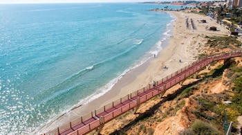 Vista aérea de playa de arena con mar azul, paseo elevado de color rojizo sobre un acantilado, bañistas, sombrillas y edificios en la costa.