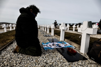 Un hombre arrodillado junto a hileras de cruces blancas, una bandera argentina y una boina roja sobre una placa de mármol negro