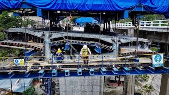 Vista aérea de trabajadores con cascos y arneses en la estructura de un puente en construcción, con andamios, vigas metálicas, hormigón y lonas azules