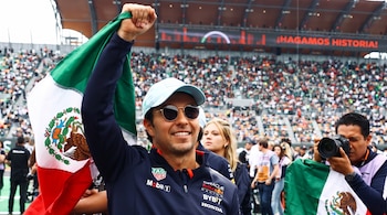 Un hombre con gorra, gafas de sol y ropa deportiva sostiene en alto una bandera de México frente a fotógrafos y gradas llenas de gente.