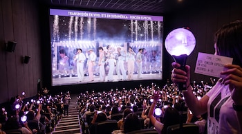 Público en una sala de cine llena sostiene bastones luminosos mientras observa en pantalla gigante un concierto del grupo musical BTS.