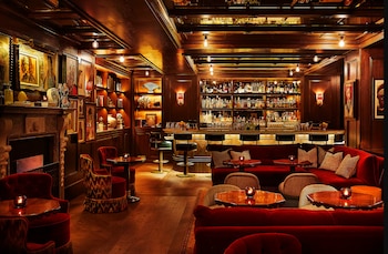 Interior de un bar de lujo con paredes y techos de madera oscura, una barra larga con estantes de bebidas y sofás y sillas de terciopelo rojo