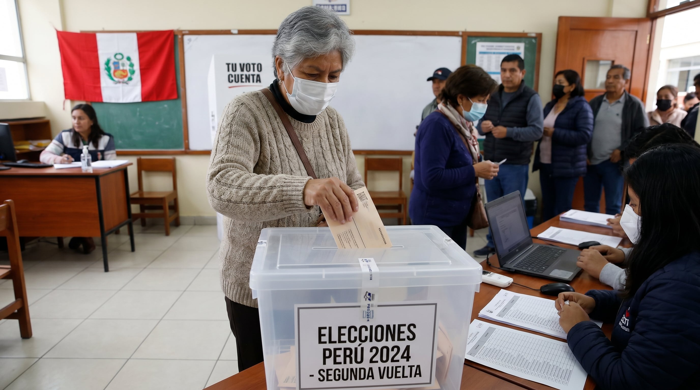 Una votante deposita su voto en una urna transparente durante la segunda vuelta de las Elecciones Generales Perú 2024, en un centro de votación con la bandera peruana de fondo. (Imagen Ilustrativa Infobae)