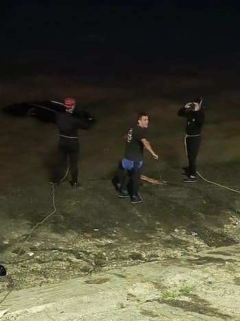Tres hombres en la oscuridad, uno con casco y lo que parece una tabla, y los otros con cuerdas, sobre un terreno desigual