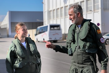 Hombre con barba y mujer joven con trajes de vuelo verdes y chalecos. El hombre señala a la mujer sonriente. Al fondo, edificios blancos y un autobús