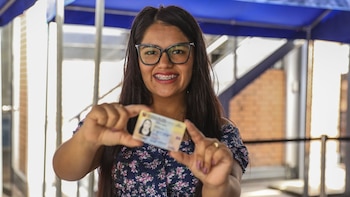 Mujer joven con gafas y frenillos sonríe mientras muestra al frente un Documento Nacional de Identidad. El fondo es un espacio público difuminado
