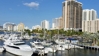 Marina con muchos barcos y yates blancos amarrados. Al fondo, edificios altos de apartamentos y oficinas, palmeras y un cielo azul despejado