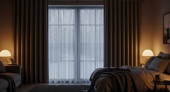 Vista de un dormitorio con cortinas térmicas marrones cubriendo las ventanas, detrás de las cuales se ven cortinas traslúcidas blancas y un exterior nevado. Dos lámparas de mesa iluminan un sofá y una cama.