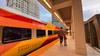 Tren Brightline rojo y amarillo en el andén de una estación. Un pasajero con equipaje camina hacia el tren. Edificios urbanos y cielo nublado visibles