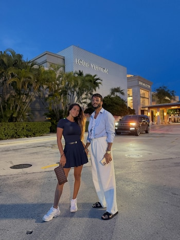 Una mujer vestida de azul y un hombre con camisa azul y pantalón blanco posan de la mano frente a un edificio con el letrero de Louis Vuitton