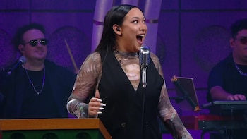 Mujer con chaleco negro y blusa estampada, boca abierta, ante un micrófono en un escenario púrpura. Dos músicos, uno con batería y otro con teclado, detrás