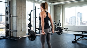 Una mujer con el pelo recogido, camiseta negra y pantalón gris, sostiene una barra de pesas en un gimnasio con suelo negro, máquinas y grandes ventanas.