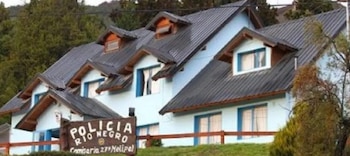 Un edificio de paredes celestes y tejado oscuro a dos aguas con buhardillas. Al frente, un cartel de madera indica 'POLICIA RIO NEGRO Comisaria 27º Melipal'