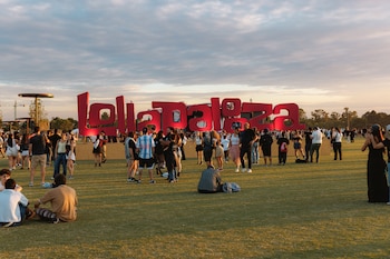 Vista panorámica de un campo de césped verde con una gran cantidad de personas de pie y sentadas frente a un letrero rojo brillante que dice 'Lollapalooza'