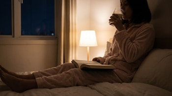 Persona en cama con pijama, bebiendo leche de un vaso y leyendo un libro. Una lámpara de noche ilumina la escena junto a una ventana oscura.