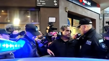 Varios policías y un hombre en el centro, rodeados de cámaras y micrófonos. Luces azules de vehículo policial en primer plano. Cartel de calle "N. Alem"