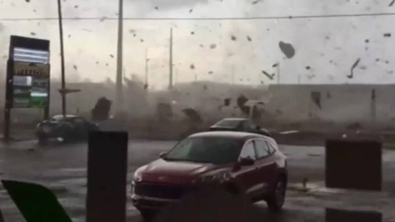 Un SUV rojo se ve en primer plano mientras escombros y polvo vuelan por el aire, reflejando la devastación causada por un fuerte tornado en una zona urbana.