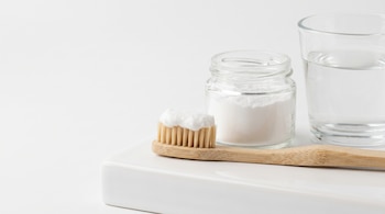 Un cepillo de dientes de bambú con pasta blanca en las cerdas, un frasco de vidrio con polvo blanco y un vaso de agua en una superficie blanca.
