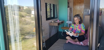 Mujer de cabello rizado y gris sentada en un sofá dentro de una mini casa con paredes de madera clara, leyendo un libro frente a una ventana con vista a un paisaje verde